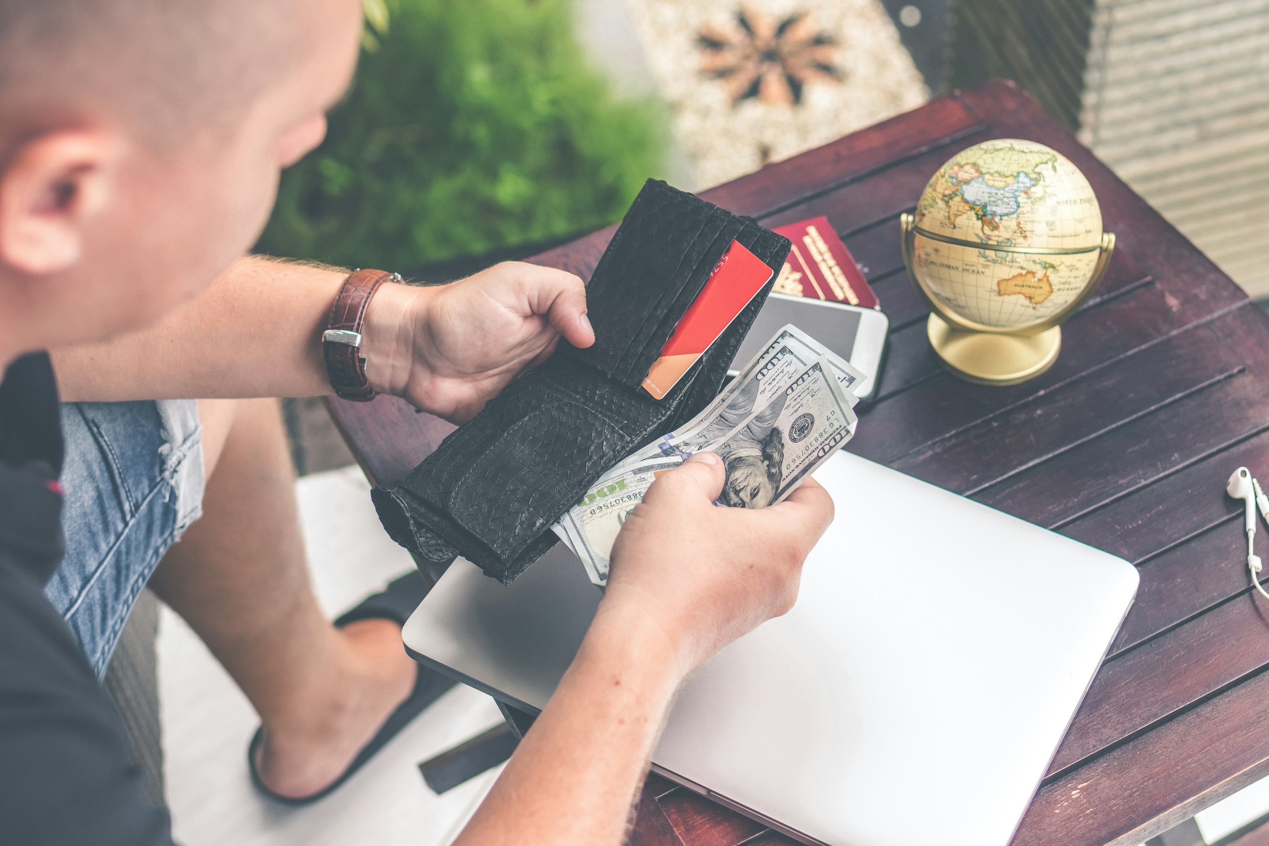 A person holding a black wallet, checking cash and credit cards, while sitting at a table with a laptop and a globe.